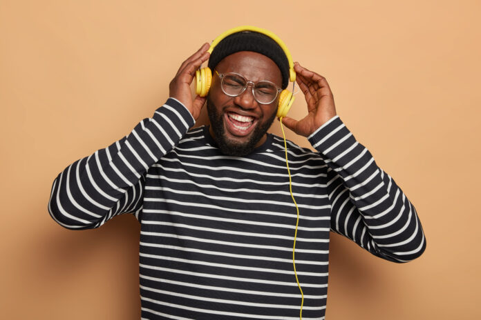 Horizontal shot of overjoyed bearded man keeps hands on headphones, wears striped jumper, listens audio track during leisure time, stands over brown background. People, music, lifestyle concept